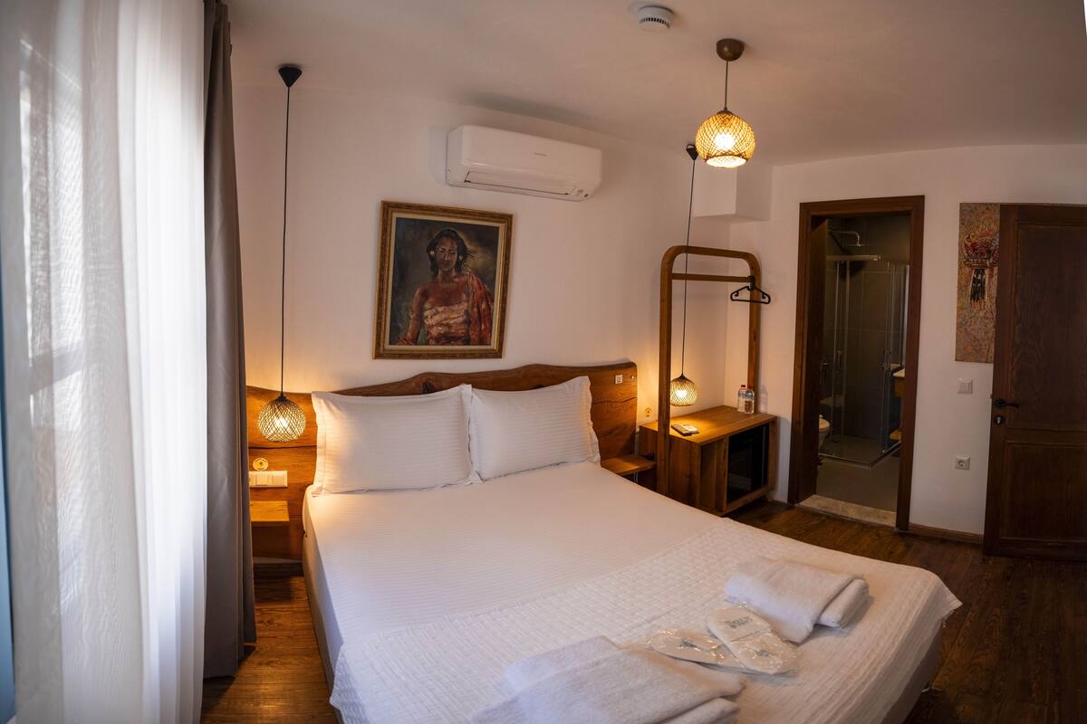 A neatly made bed with white linens, a bedside table with a lamp, a painting on the wall, and an air conditioning unit.