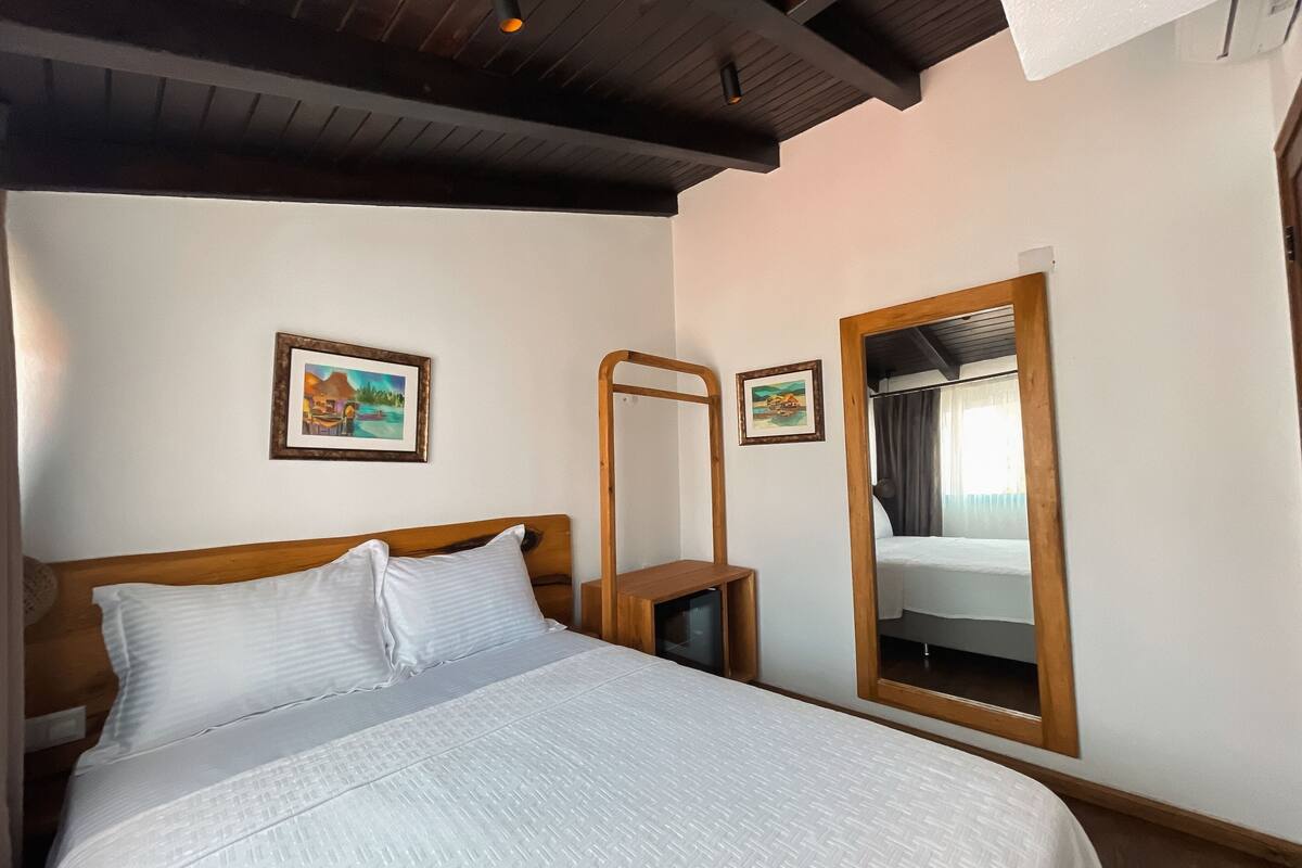 A neatly made bed with white linens, a wooden headboard, and a wooden nightstand beside it. There are two framed pictures on the wall, one above and one below the bed. A mirror with a wooden frame is mounted on the wall to the right of the bed.
