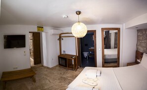 A bedroom with a bed, a wooden bench, a hanging light fixture, a TV, and a bathroom visible through an open door. - Casa Alegre Alaçatı (Cesme)
