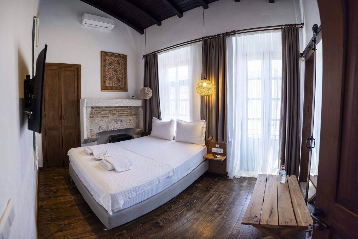 A bedroom with a large bed, wooden floor, a TV mounted on the wall, and a wooden table with a bottle.