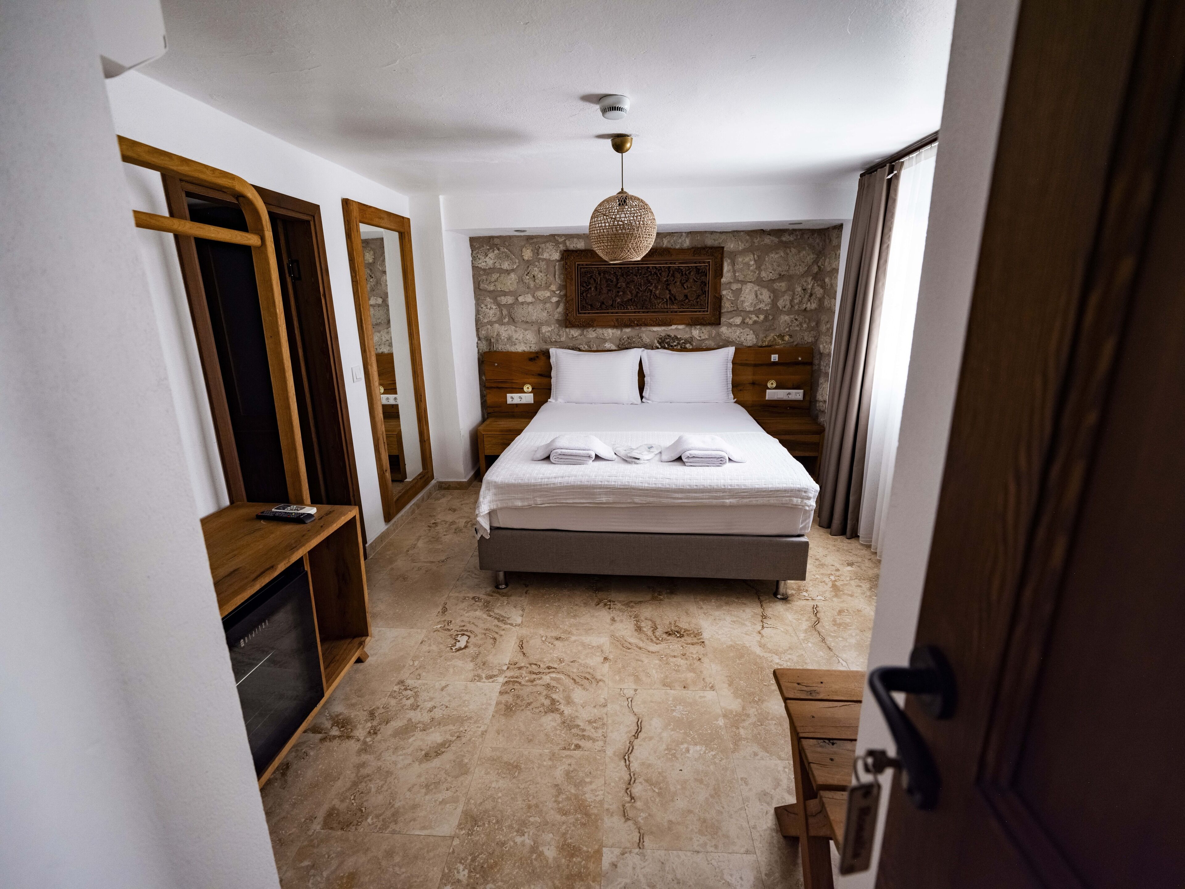 A bedroom with a stone wall, a bed with white linens, wooden headboard, bedside tables, and a hanging light fixture.