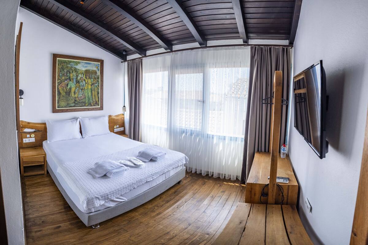 A bedroom with a large bed, wooden flooring, a painting on the wall, a TV mounted on the wall, and a window with sheer curtains.