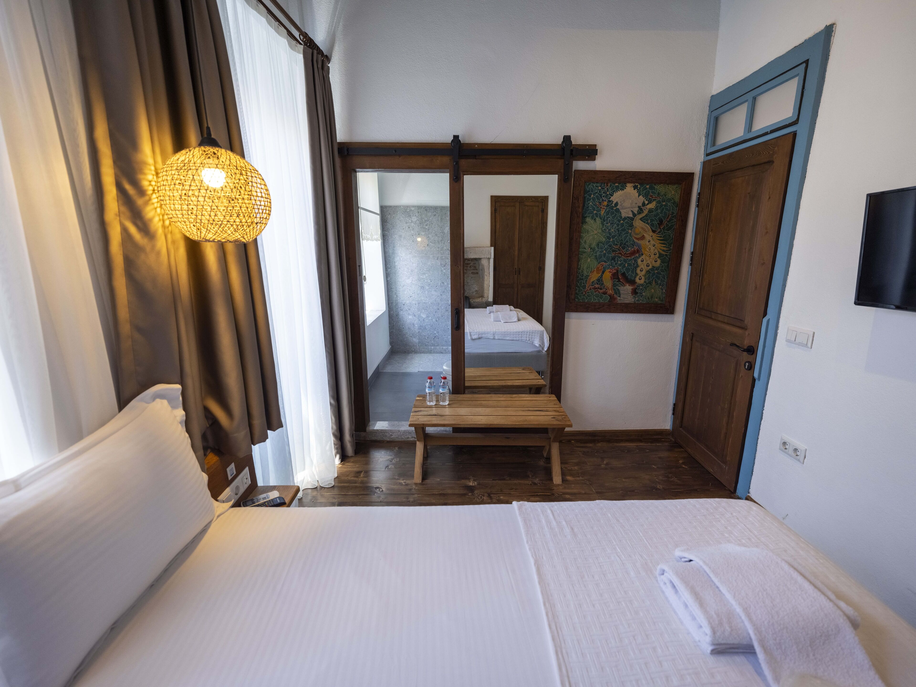 Een hotelkamer met een bed, een houten bankje, een tafel met waterflesjes, een aan de muur bevestigde televisie en een deur naar een badkamer.