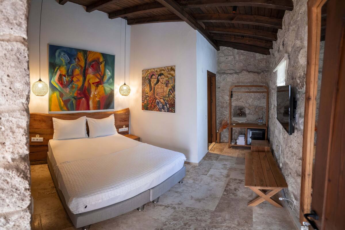 A bedroom with a large bed, wooden headboard, two paintings, a stone wall, a stone bench, and a television mounted on the wall.