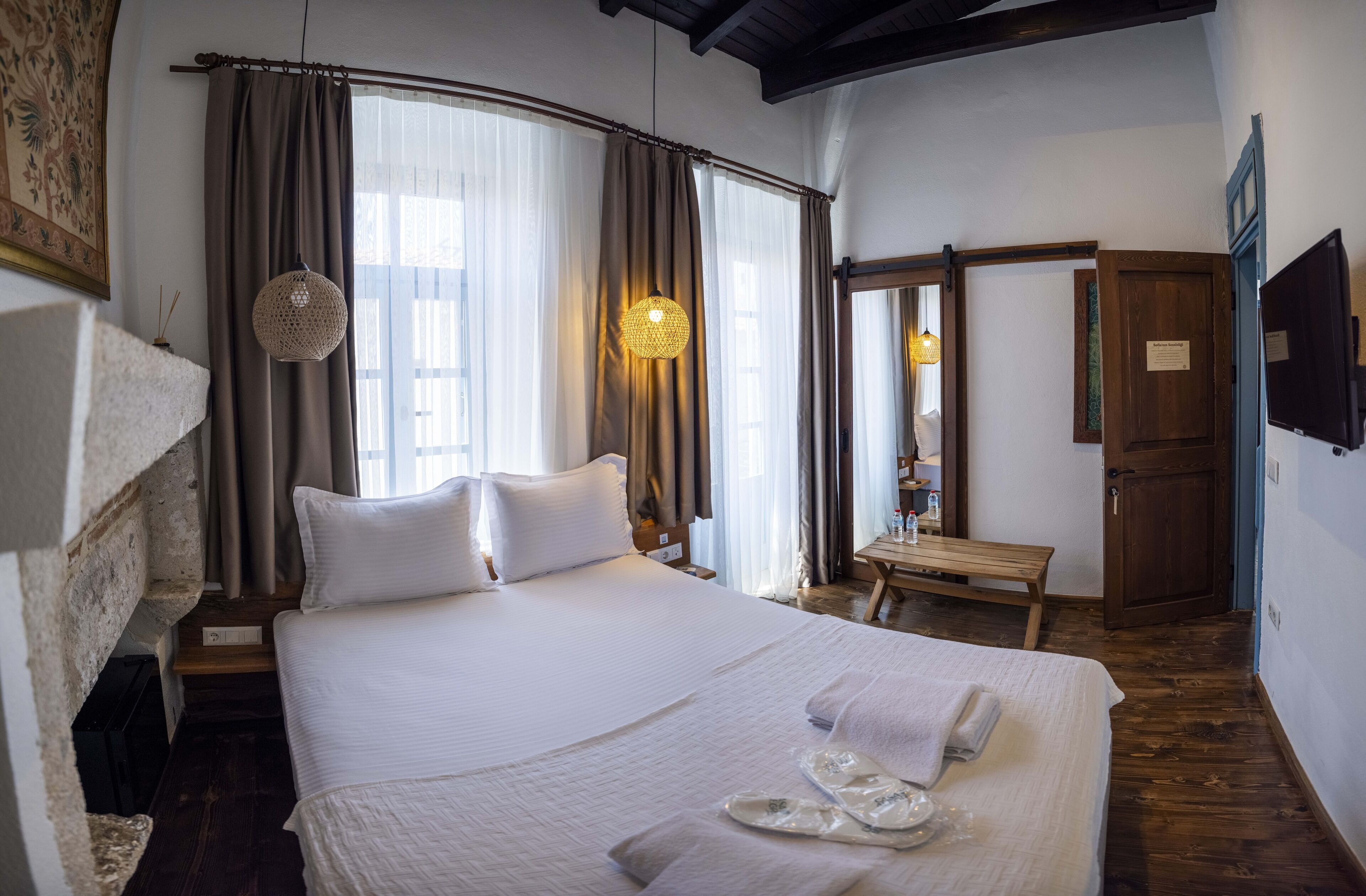 Una cama bien hecha con sábanas blancas, un banco de madera y una lámpara colgante en una habitación con piso de madera y pared de piedra.