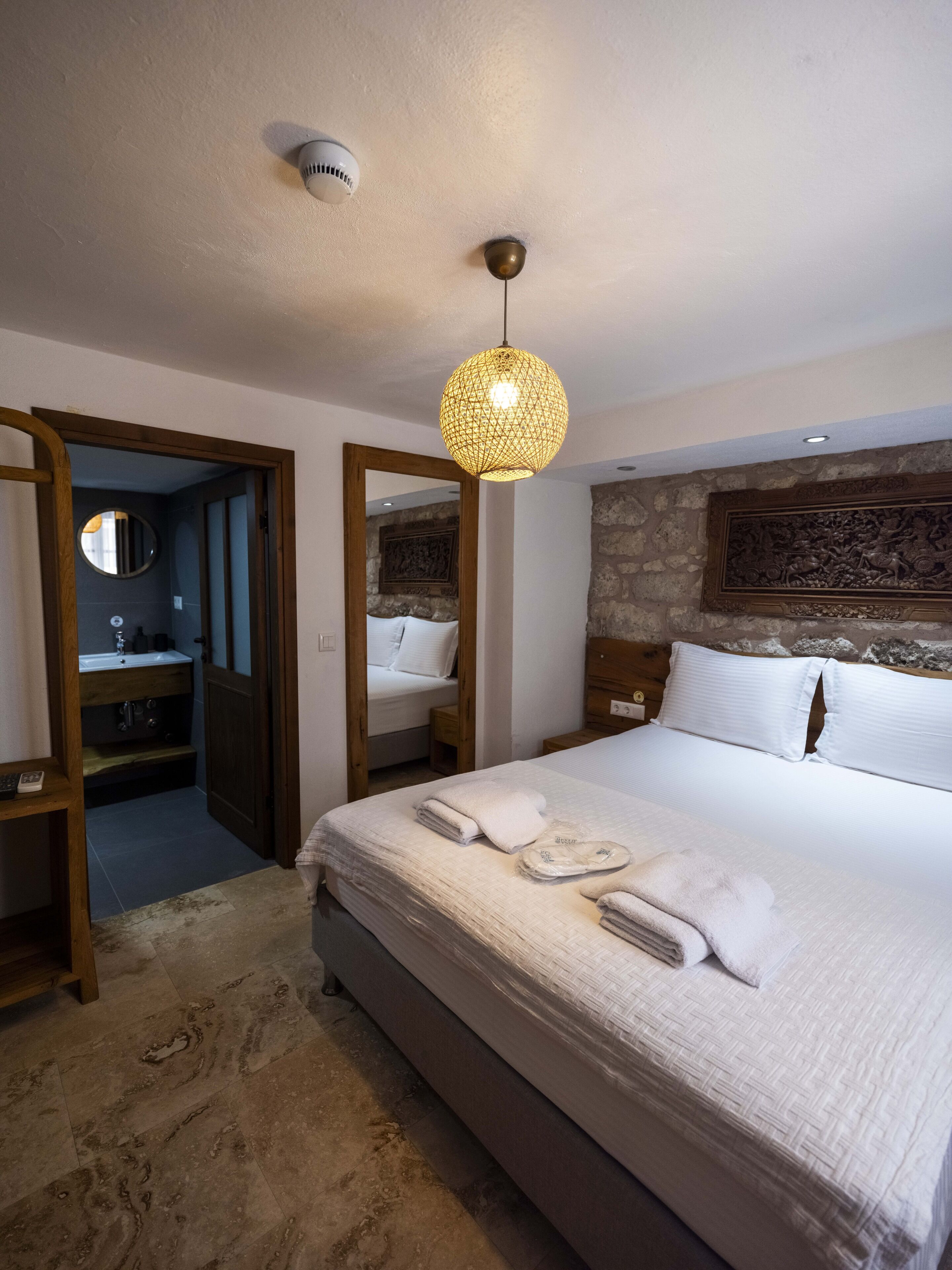 A hotel room with a large bed, a wooden wardrobe, a round mirror, a stone wall, and a ceiling light.