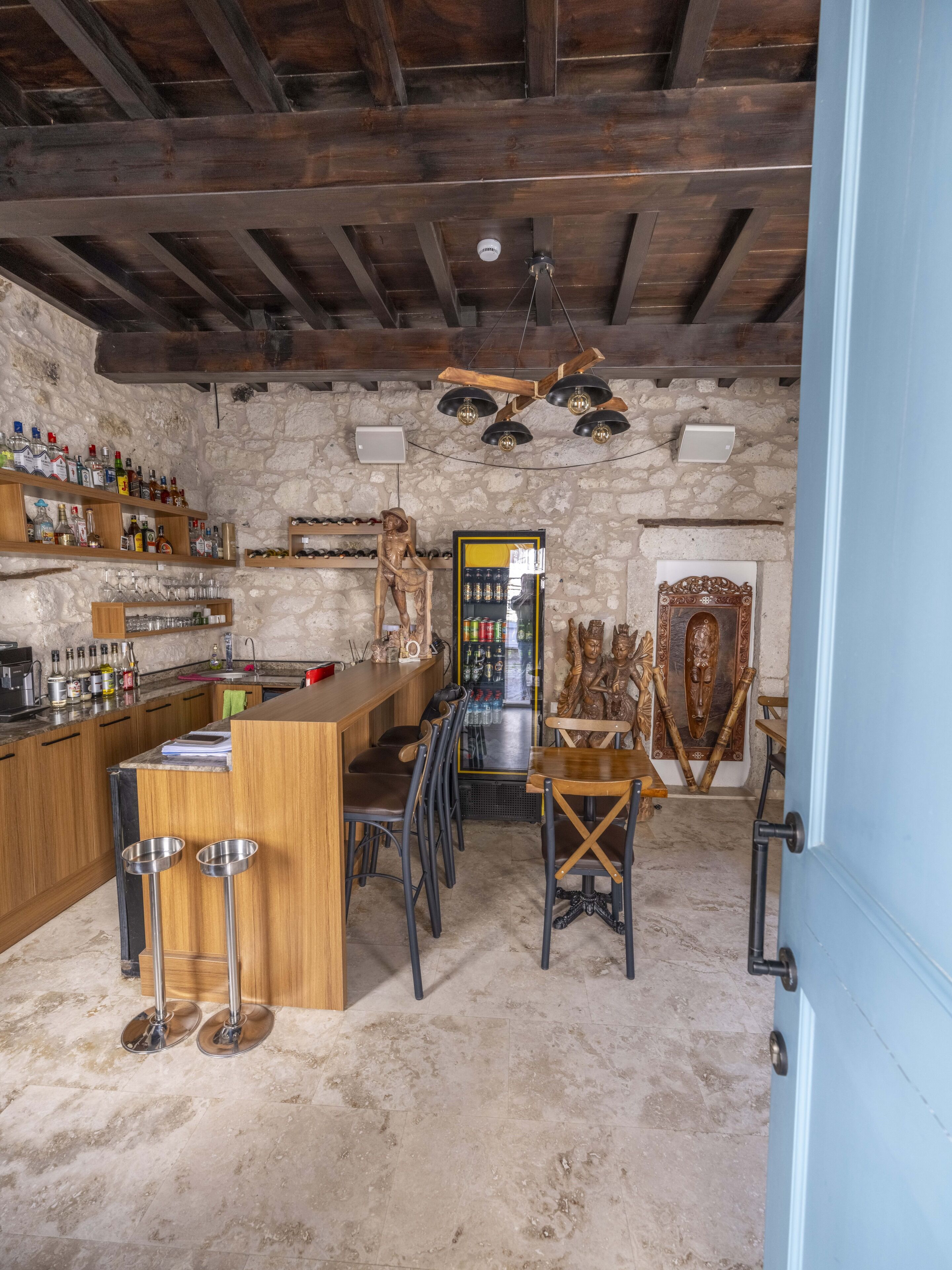 Un rústico área de bar con estantes de madera, una barra y taburetes. Se observan máscaras decorativas y una pared de piedra al fondo.