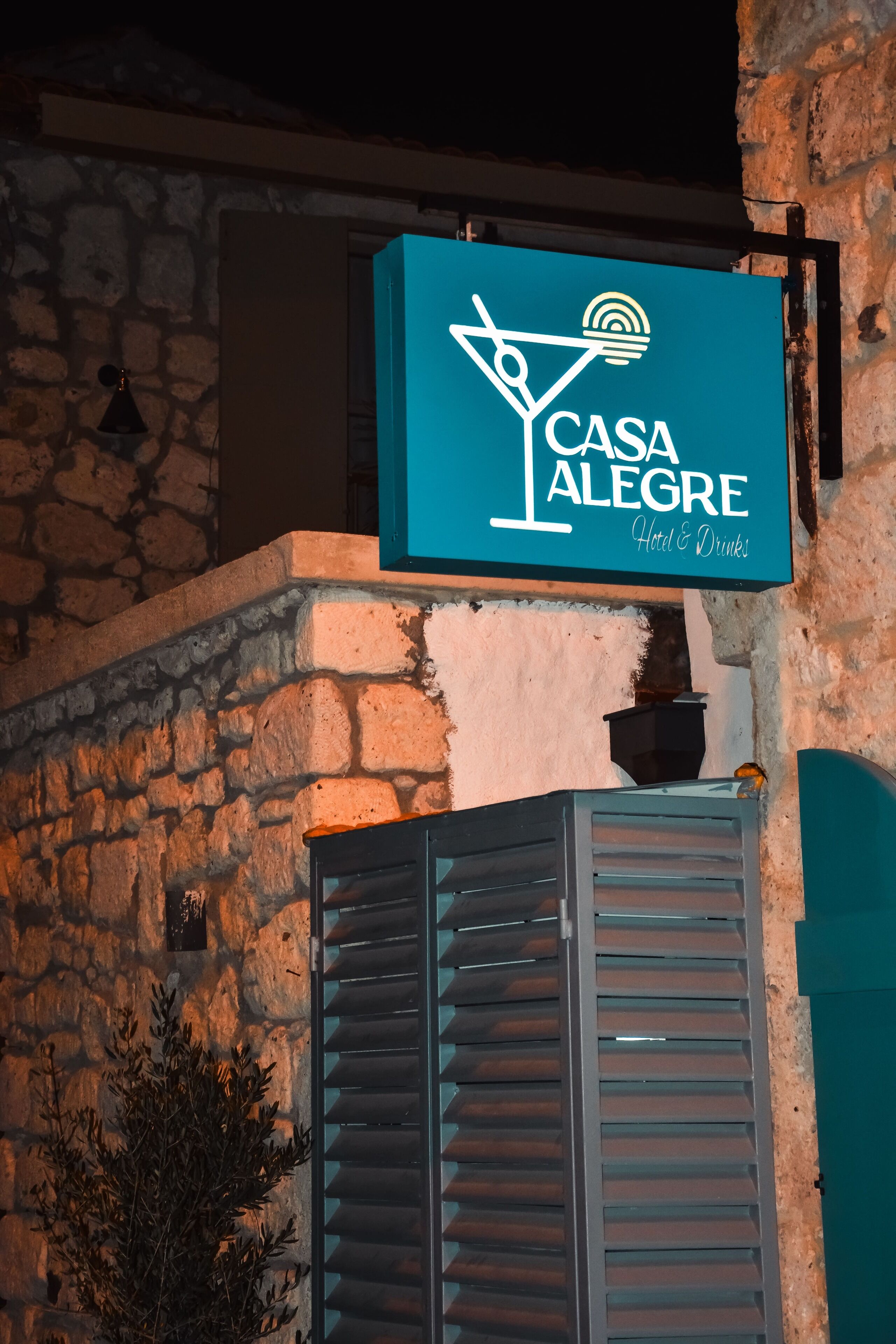 Een bord met 'Casa Alegre Hotel & Drinks', met een cocktailglas en een zonnetje, bevestigd aan een stenen muur met luiken.