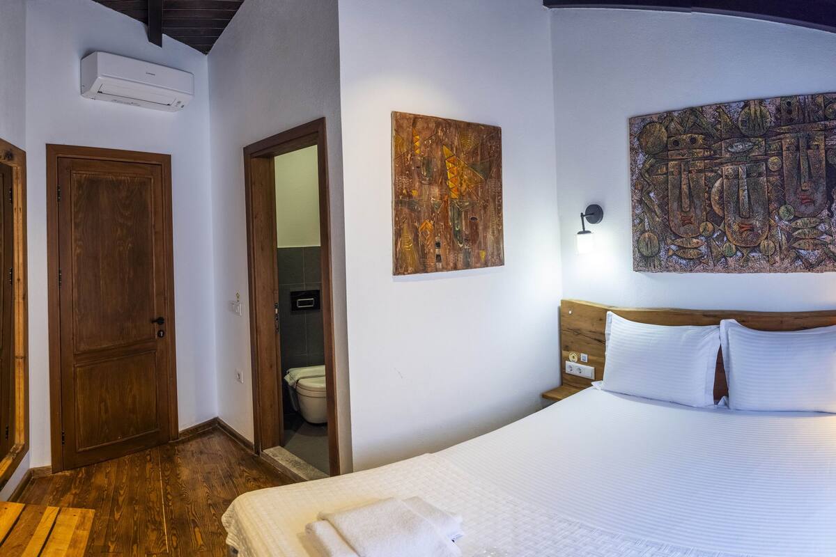 A hotel room with a wooden floor, a bed with white bedding, a mounted TV, wooden doors, and artwork on the walls.