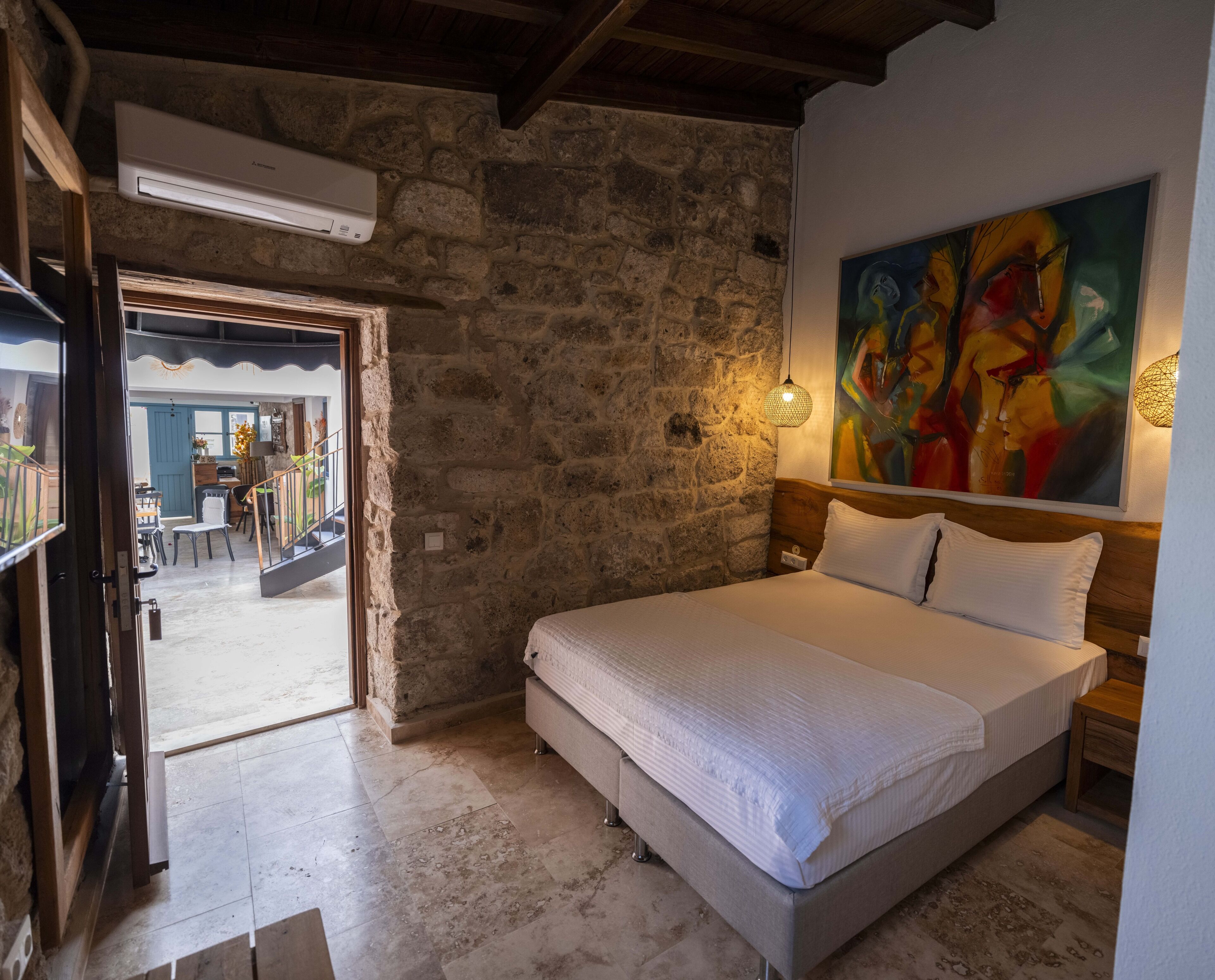 A bedroom with a stone wall, a bed with white linens, a painting on the wall, and a hanging light fixture.