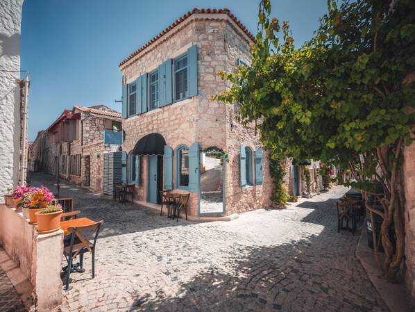 A stone building with blue shutters and a café area with outdoor seating. - Casa Alegre Alaçatı (Cesme)