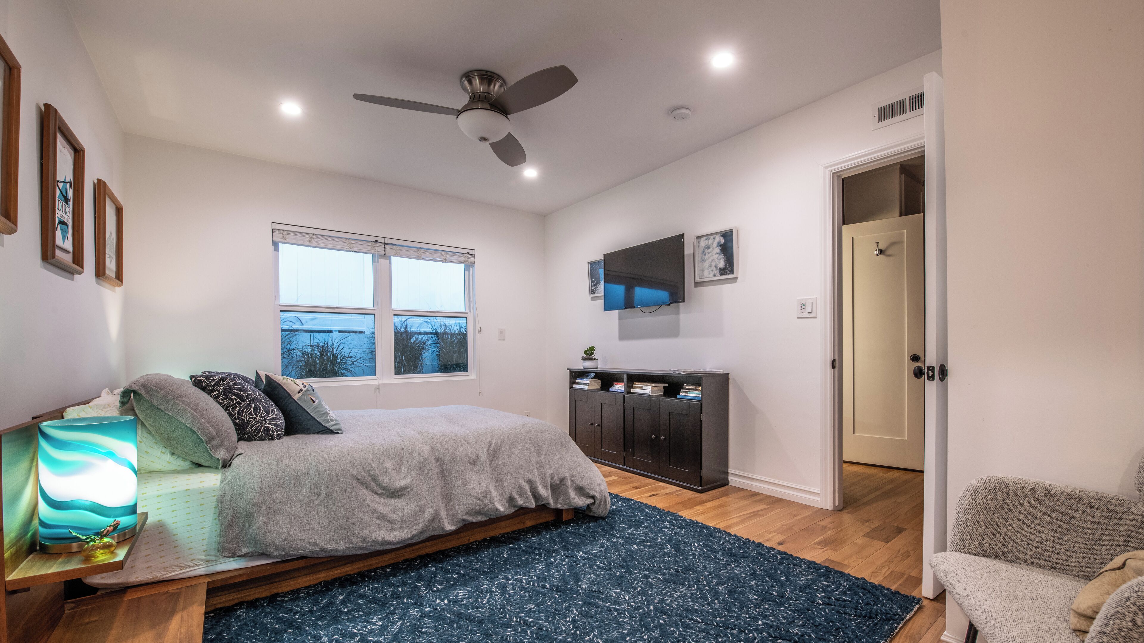 Ein Schlafzimmer mit Bett, Schreibtisch, Stuhl, Fernseher, Deckenventilator und Fenster.