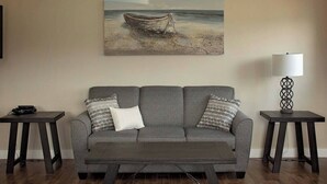 A living room with a grey sofa, two side tables, a coffee table, a painting of a boat on a beach, and a floor lamp. - Gros Morne Coastal Suites #2 (Rocky Harbour)
