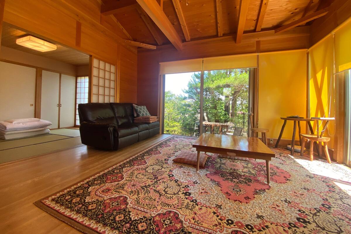 A traditional Japanese-style room with tatami flooring, a black leather sofa, a wooden table, and a large window offering a view of greenery.