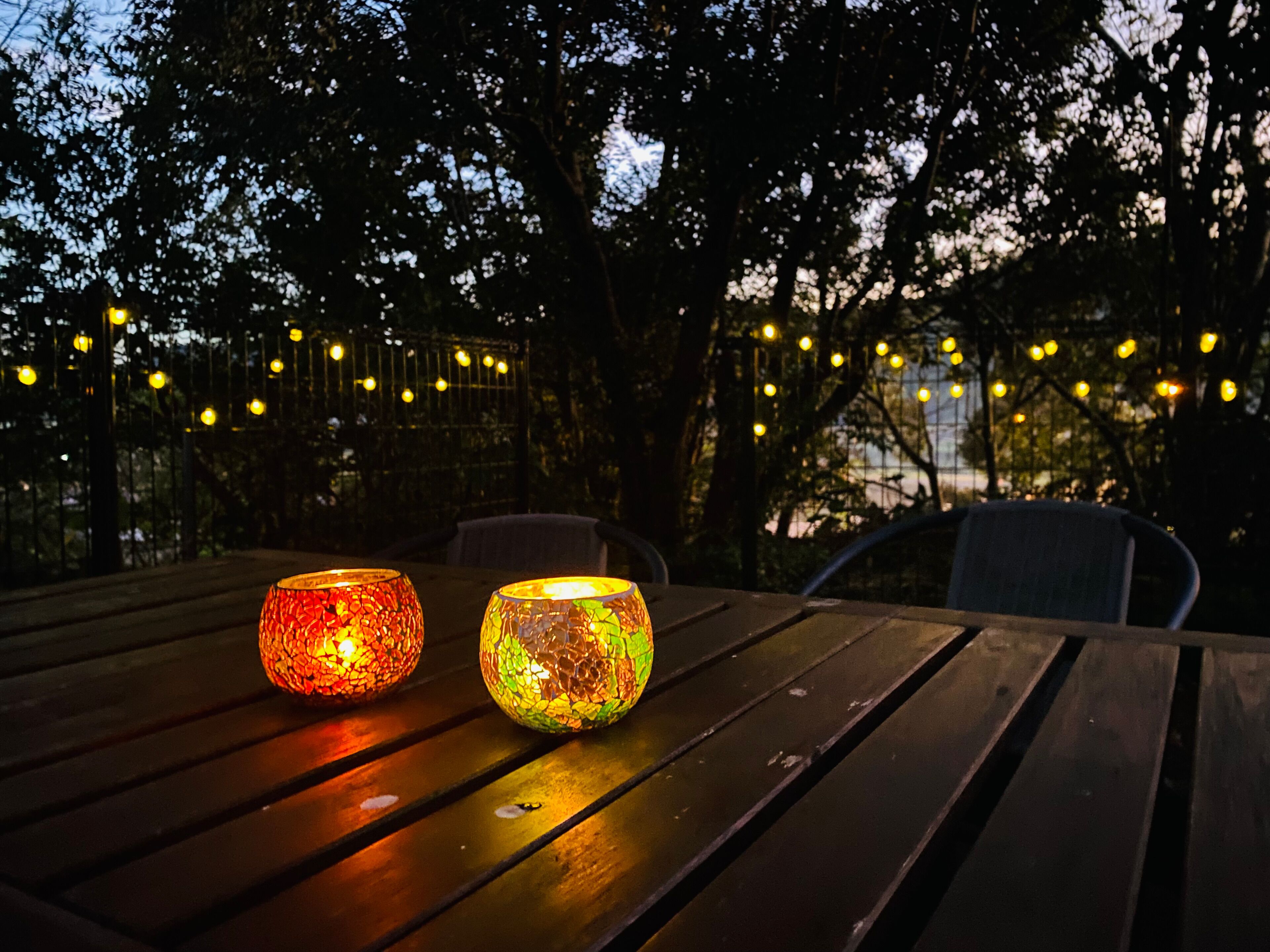 Two illuminated glass lanterns on a wooden table outdoors at dusk.