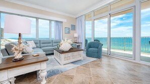 A spacious living room with a large window overlooking the ocean, a grey sofa, a wooden coffee table, and a blue armchair.