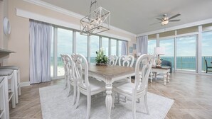 A spacious dining area with a wooden table and white chairs, large windows offering a view of the ocean, and a ceiling fan.