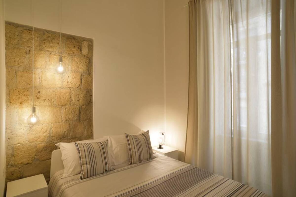 A bedroom with a stone wall, a bed with striped bedding, two bedside lamps, and sheer curtains.