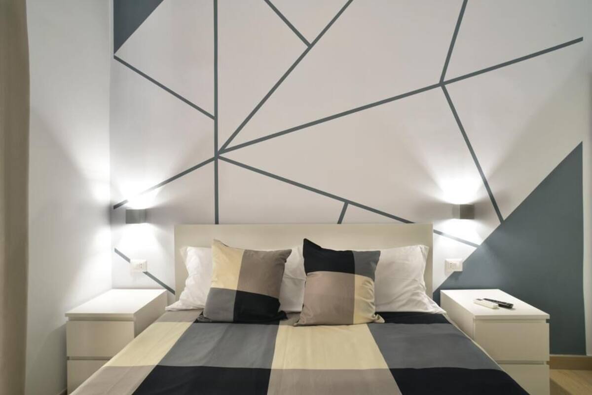 A modern bedroom with a geometric-patterned headboard, a bed with a striped bedspread, two bedside tables, and wall-mounted reading lights.