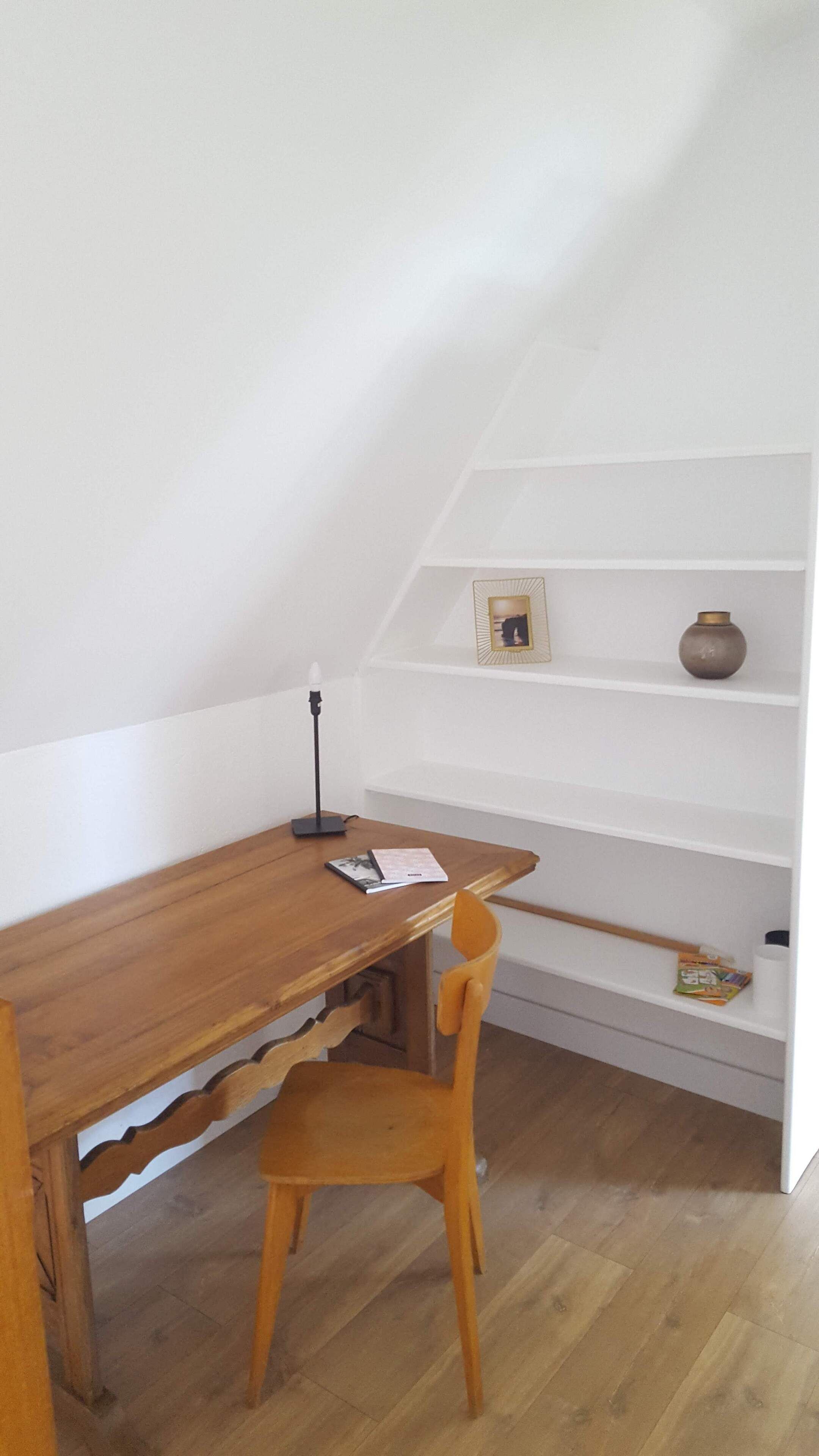 A small, cozy room with a wooden desk, a chair, a lamp, and a shelf with a vase and a framed picture.