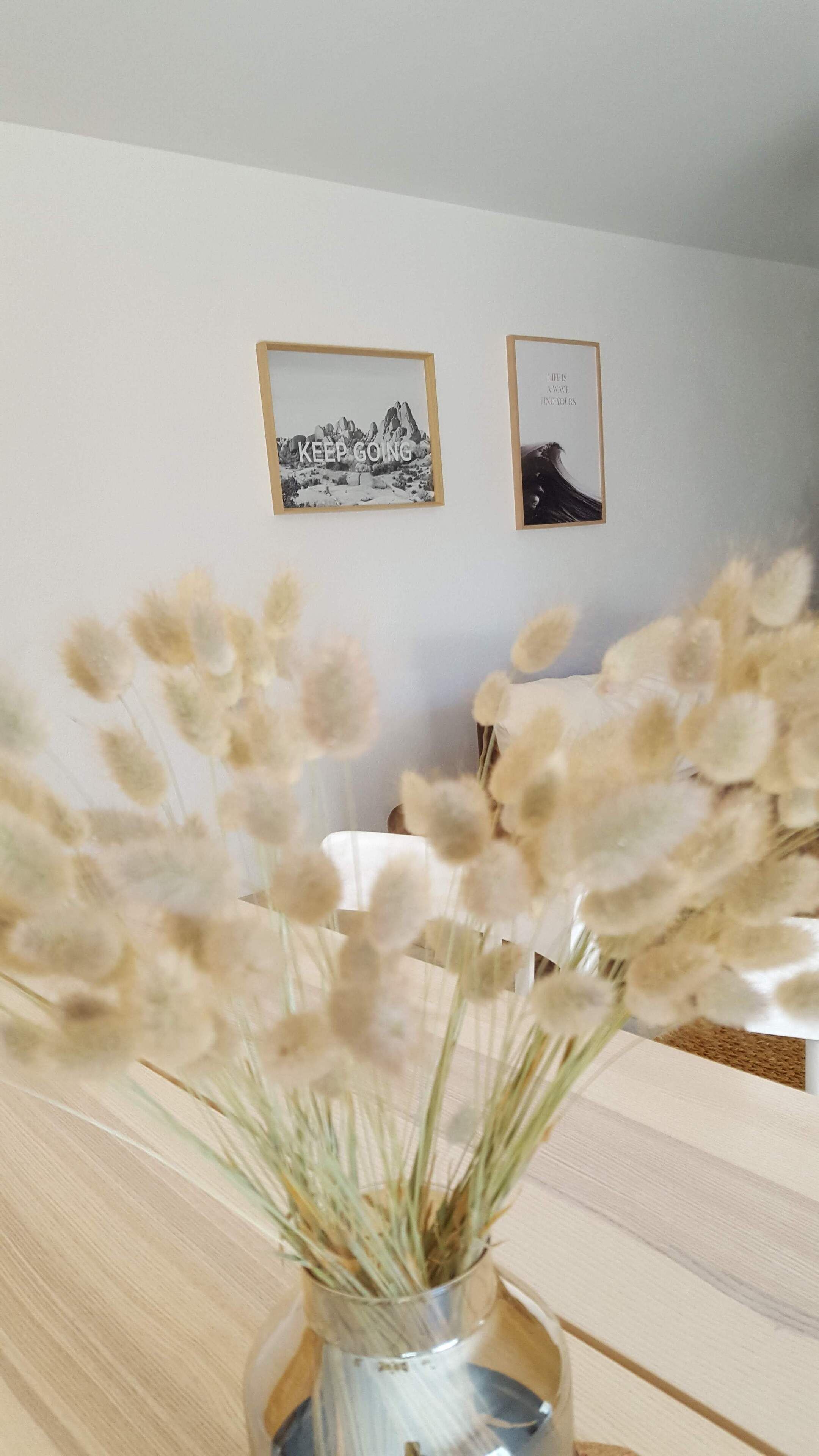 Ett rum med ett träbord, en vas med torkade blommor och två inramade tavlor på väggen.