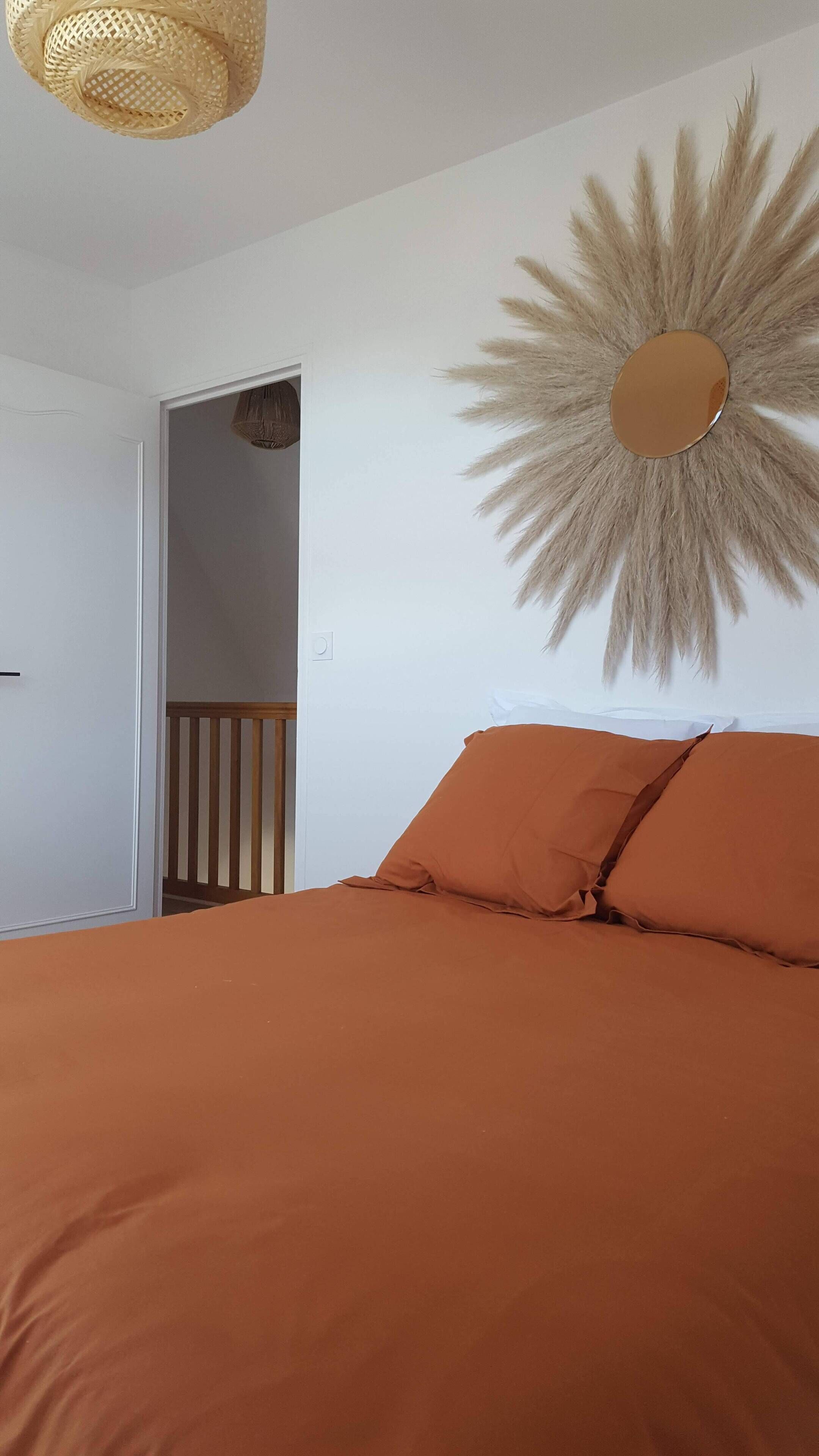 A bedroom with an orange bedspread, a wooden headboard, a mirror with a sunburst design, and a hanging light fixture.