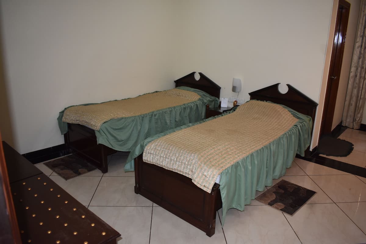 Una habitación con dos camas de madera, cada una con colcha verde y manta color beige. Entre las camas hay una mesita con cubierta estampada y una lámpara de pared.