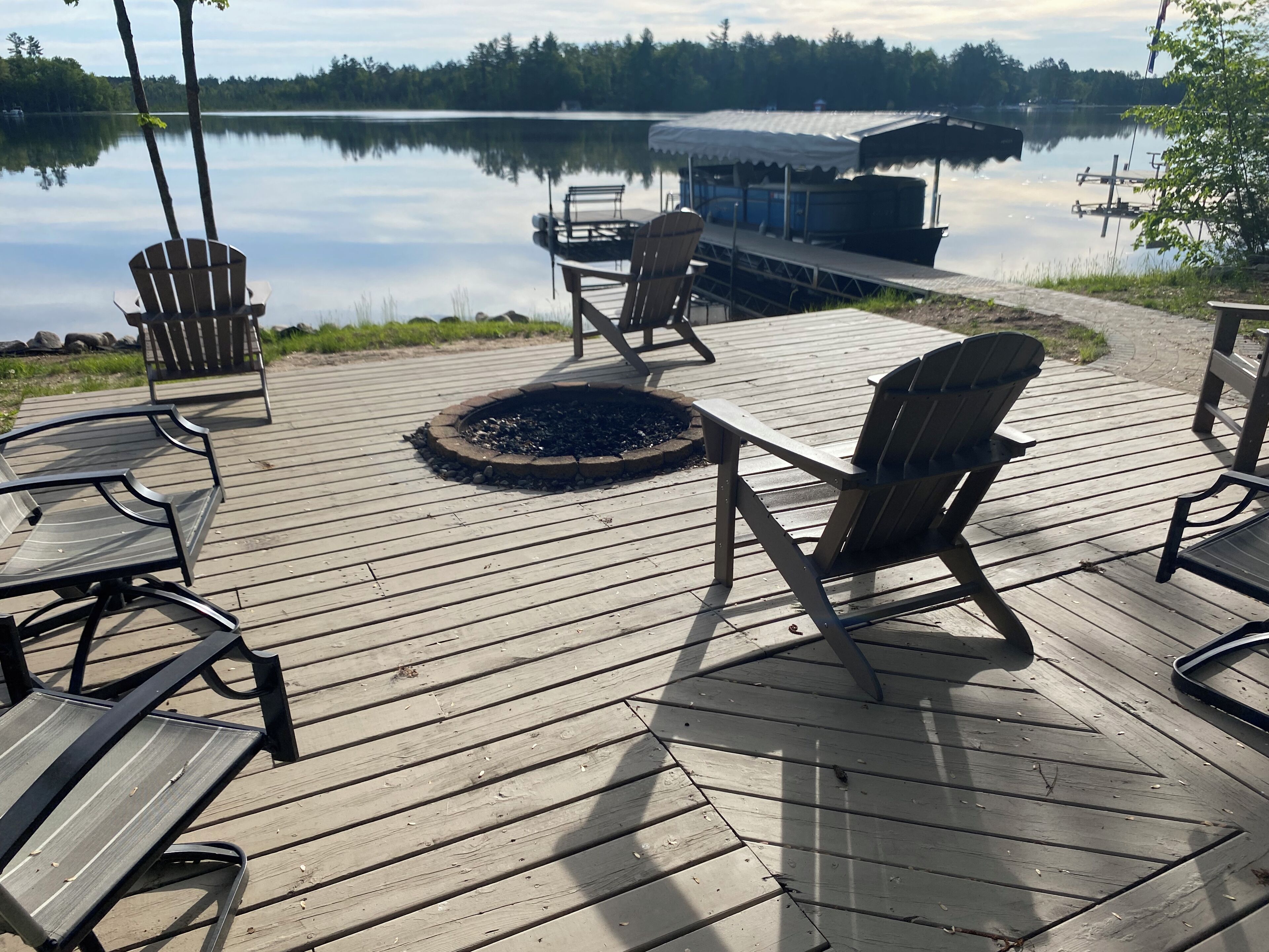A wooden deck with Adirondack chairs and a fire pit, overlooking a calm lake and dock.