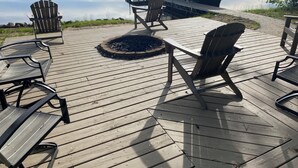 Una terraza de madera con sillas Adirondack y un hogar para fogata, con vista a un lago tranquilo y un muelle.