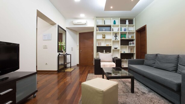 Un salon moderne avec un téléviseur à écran plat, un canapé gris, une table basse en verre et un pouf beige. Le sol est en bois, le mur est blanc et il y a une bibliothèque intégrée agrémentée de divers objets décoratifs.
