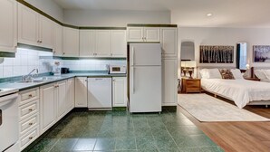 A compact kitchen with white cabinets, a refrigerator, and a microwave. Adjacent is a bedroom with a bed, nightstands, and artwork. - Welcome to “Chateau Sherbrooke's” loft: an historic gem in Downtown Montreal. (Montréal)