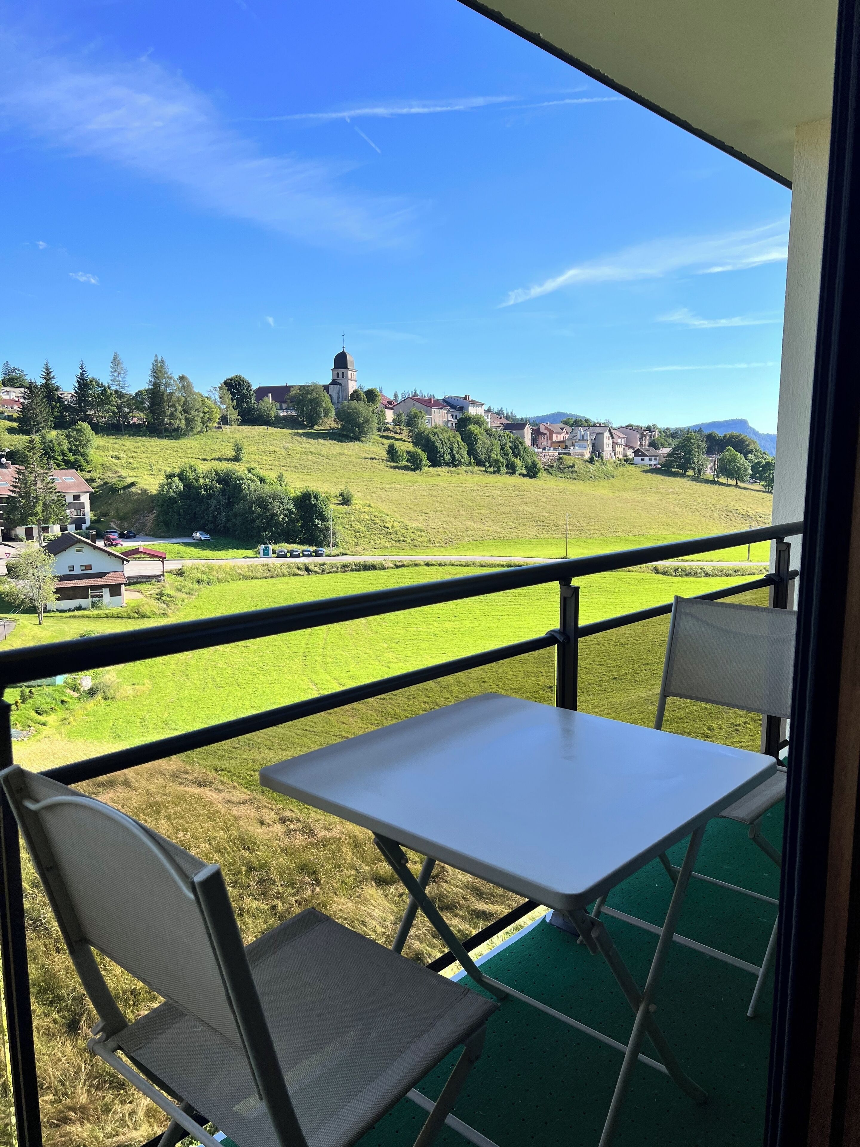 Een balkon met een tafel en stoelen, met uitzicht op een groen landschap met huizen en een kerktoren.