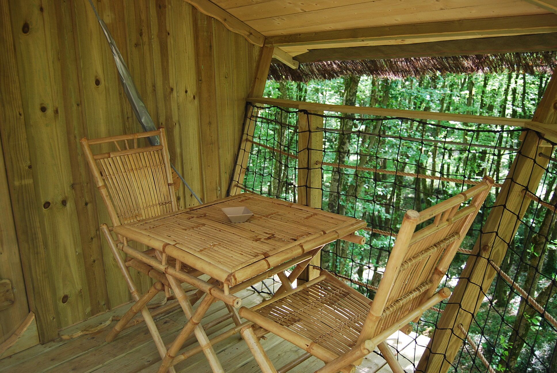 A wooden room with a table and chairs, a thatched roof, and a view of a forest.