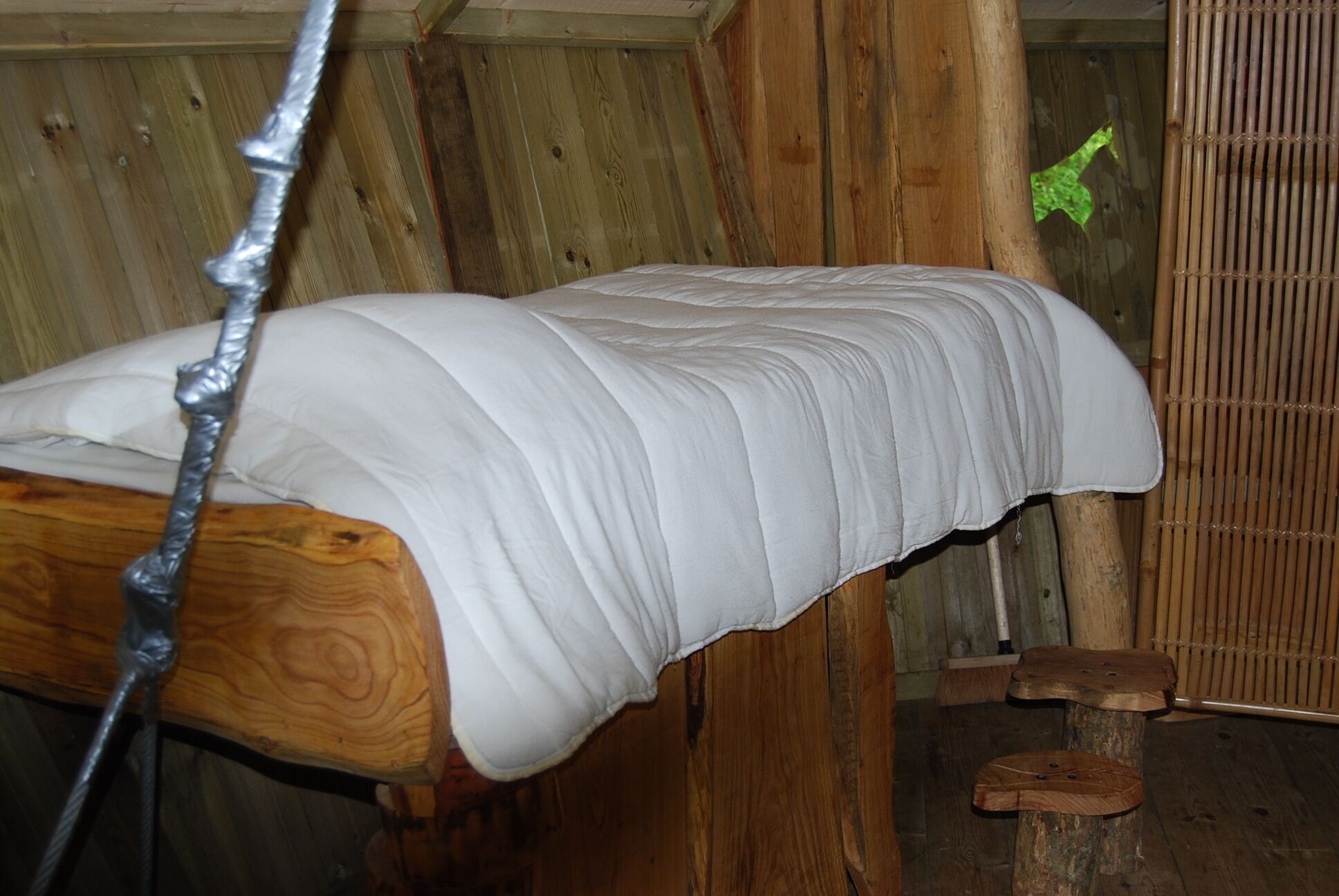 Un lit en bois avec un matelas blanc, dans une chambre rustique aux murs en bois et agrémentée d’un paravent en bambou.