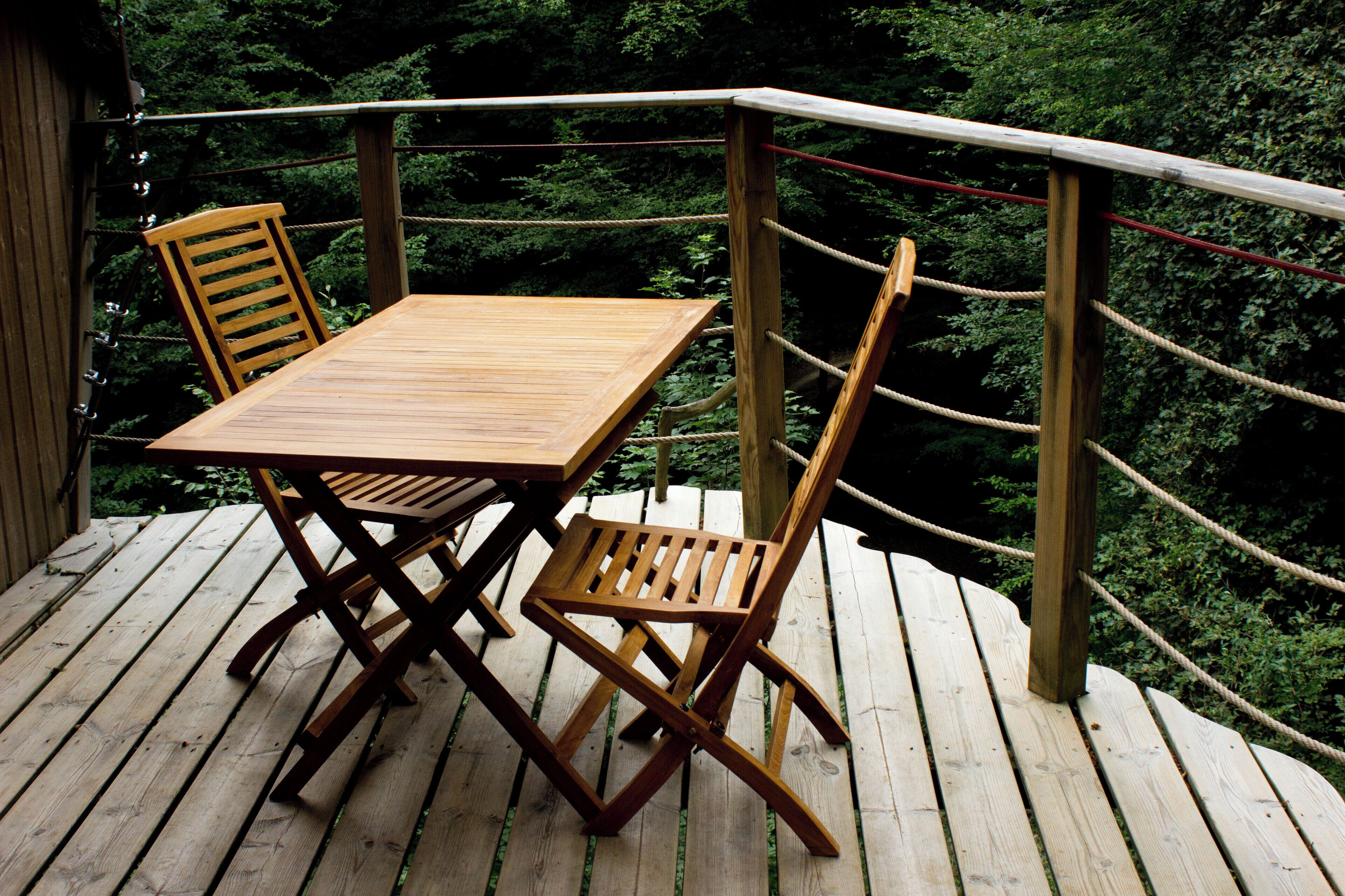 A wooden deck with a table and two chairs, surrounded by a rope barrier and dense foliage in the background.