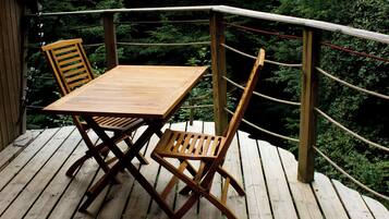 A wooden deck with a table and two chairs, surrounded by a rope barrier and dense foliage in the background.