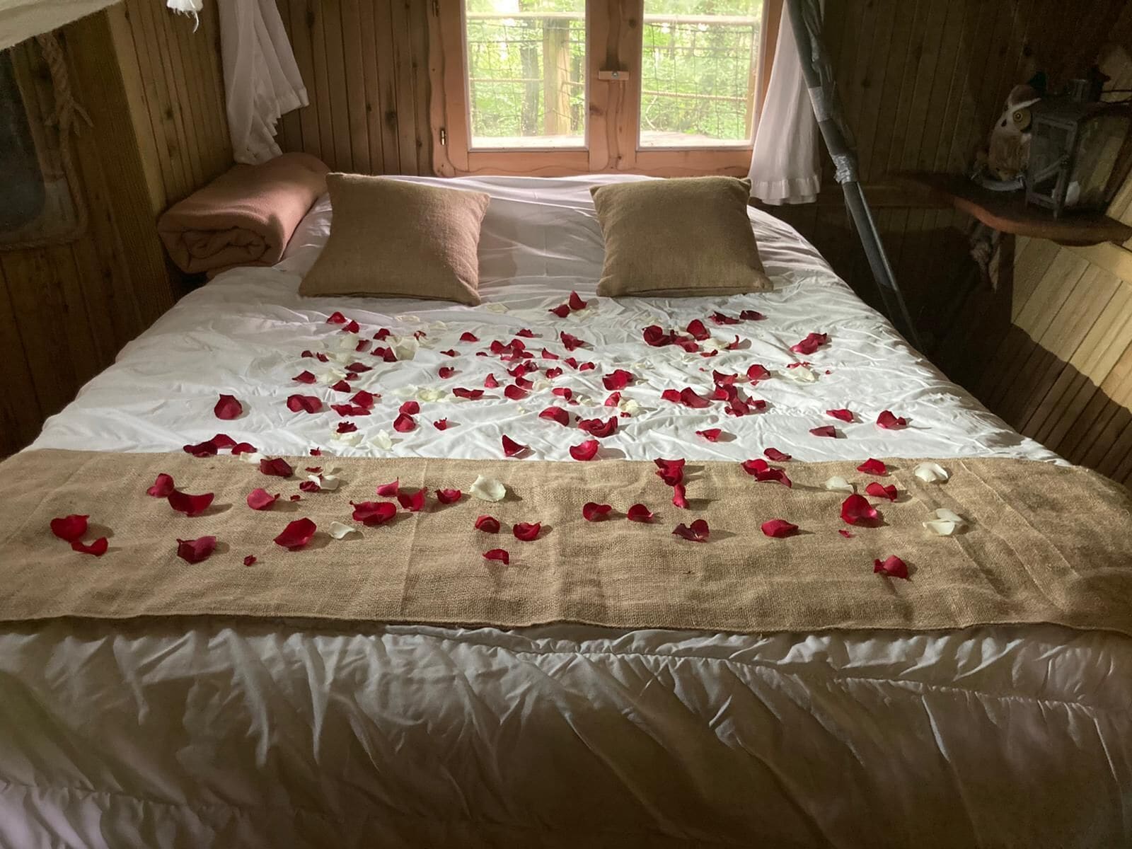Ein Zimmer aus Holz mit einem Bett, das mit weißen Leinen und Rosenblättern bezogen ist, zwei Kissen und einem Fenster mit Blick auf Grünflächen.
