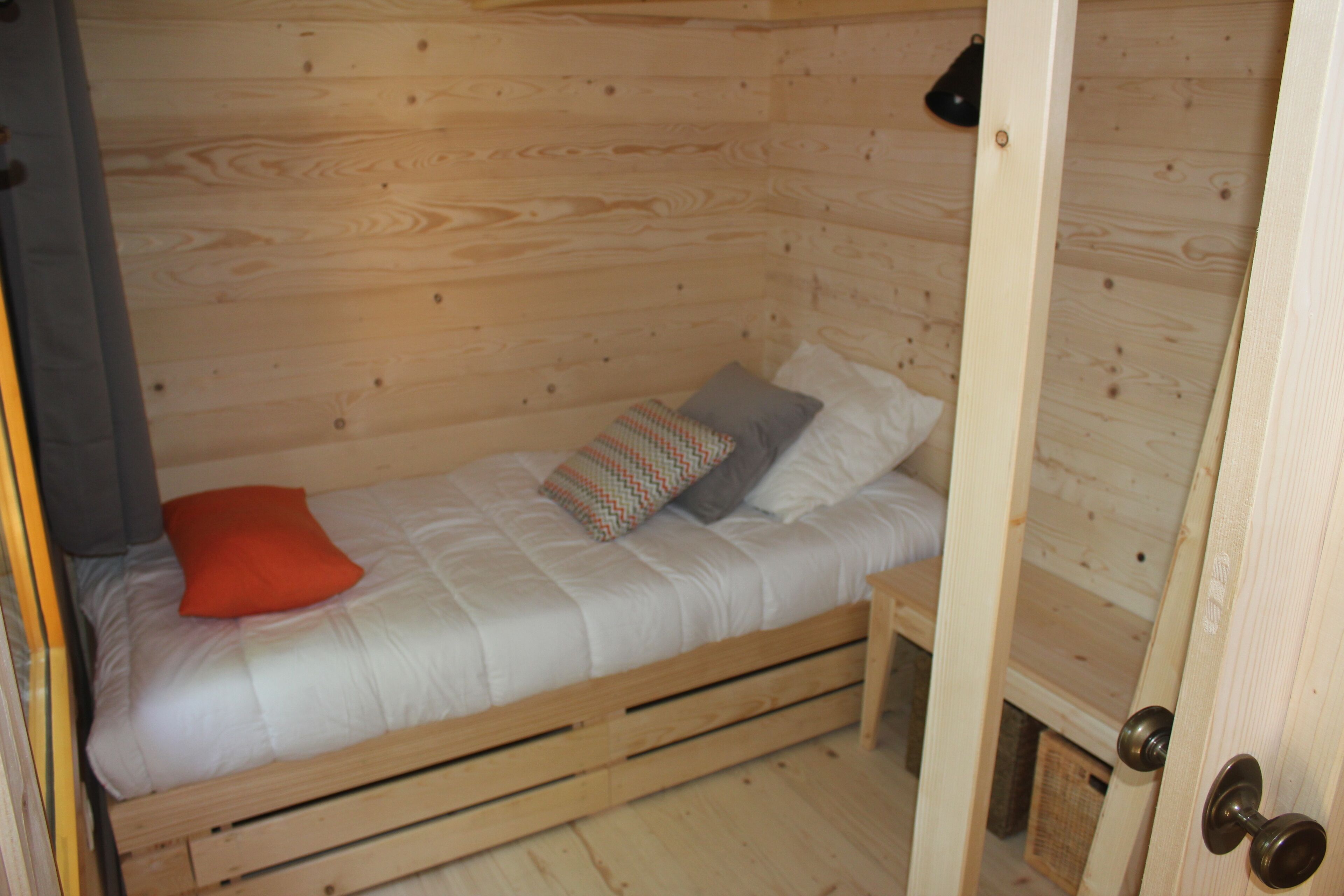 Un simple lit en bois, avec un matelas, des oreillers et une couverture, dans une pièce aux murs en bois et dotée d’une porte.