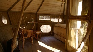 A rustic cabin interior with wooden walls, a thatched roof, a small table, a chair, and a bed. - Le Parc de la Belle (Magné)