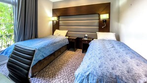 A hotel room with two single beds, a wooden headboard, bedside lamps, a small table, and a window with curtains. - LHA Paddington (London)