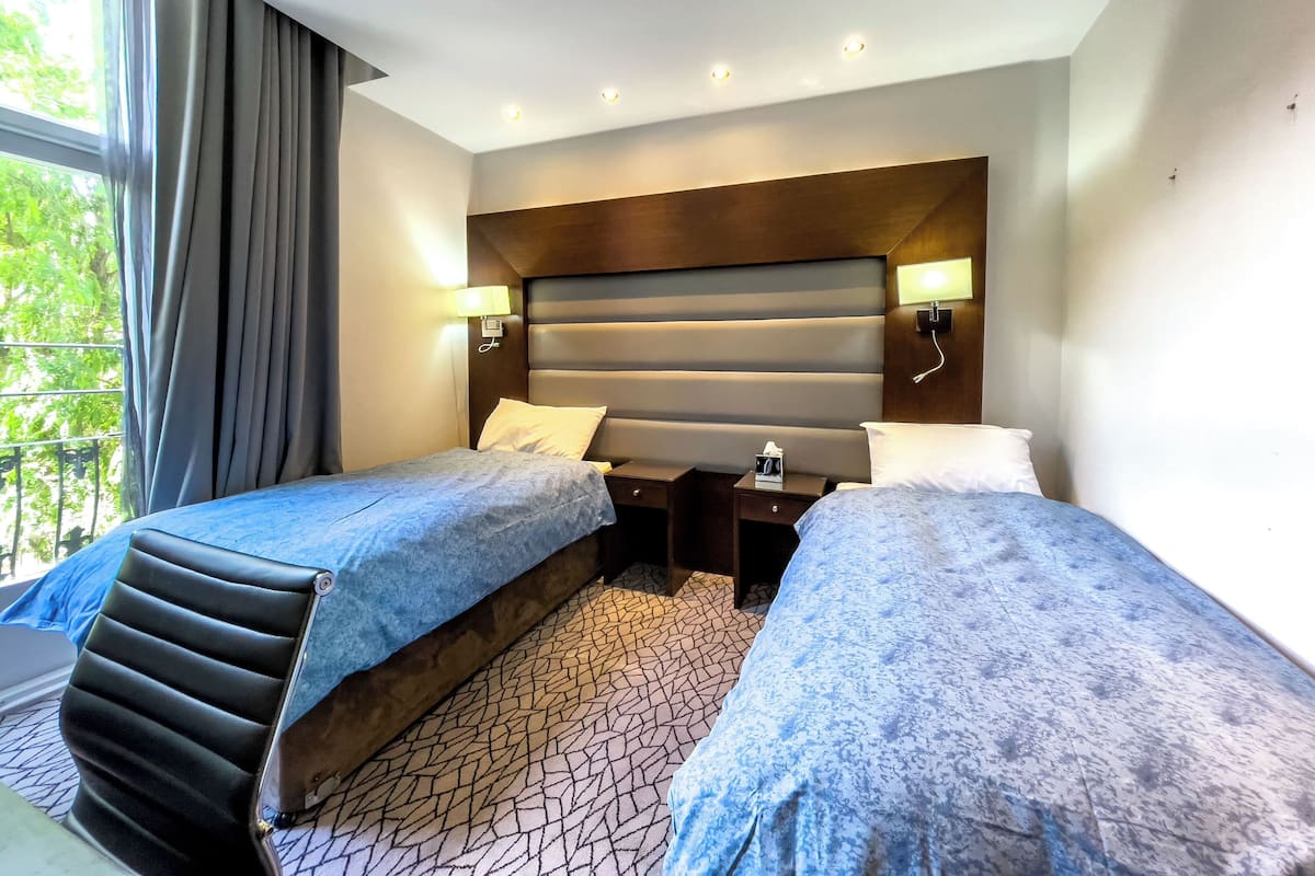 A hotel room with two single beds, a wooden headboard, bedside lamps, a small table, and a window with curtains.