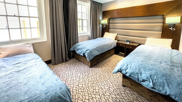 A hotel room with two beds, a large window with curtains, a headboard with built-in lighting, and a patterned carpet. - LHA Paddington (London)
