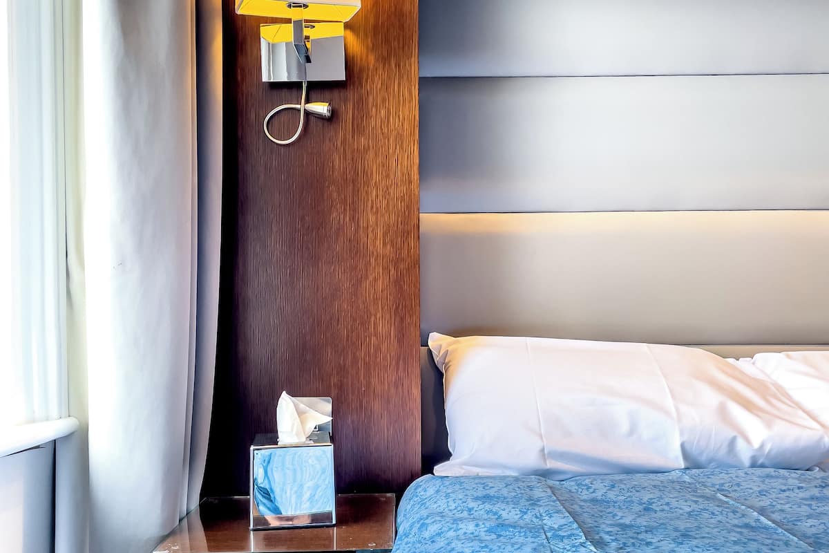A hotel room with a wooden headboard, a bedside table with a lamp, a tissue box, and a neatly made bed with white sheets.