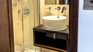 A modern bathroom with a glass-enclosed shower, a white sink, and a stainless steel trash can. - LHA Paddington (London)