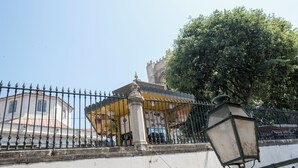 Ein historisches Gebäude mit gelber Fassade, einer weißen Mauer mit Metallzaun und einem Baum im Hintergrund.