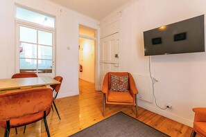 A room with a wooden floor, a dining table with chairs, a TV mounted on the wall, and a door leading to another room.