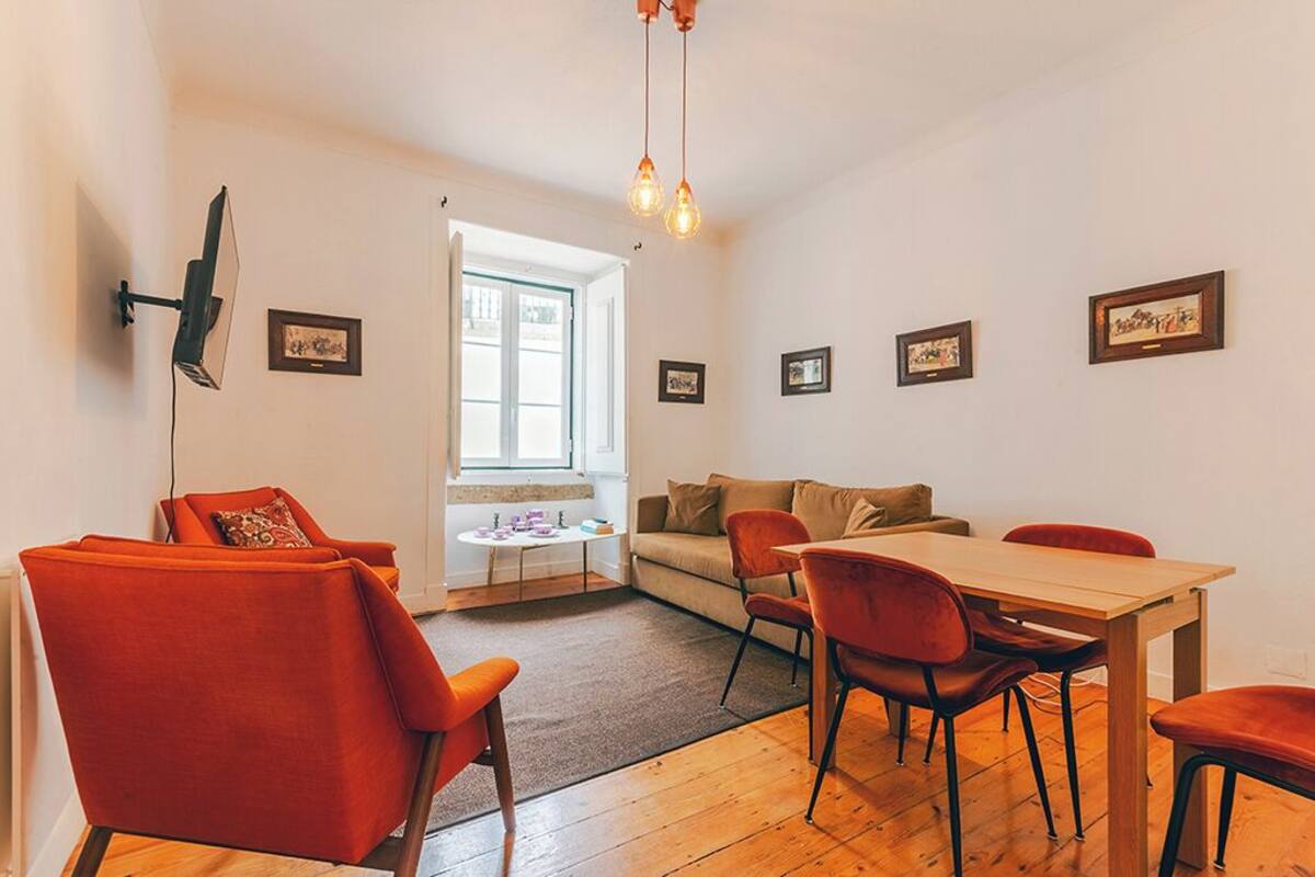 A dining area with a wooden table and red chairs, a sofa, and a TV mounted on the wall.