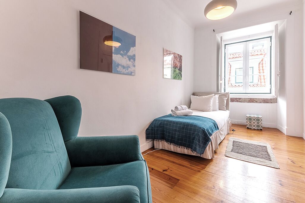 A room with a teal armchair, a bed with a blue checkered blanket, and a window with a view of buildings.