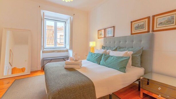 Ein Schlafzimmer mit einem Bett, einem Fenster, einer Lampe und gerahmten Bildern an der Wand.