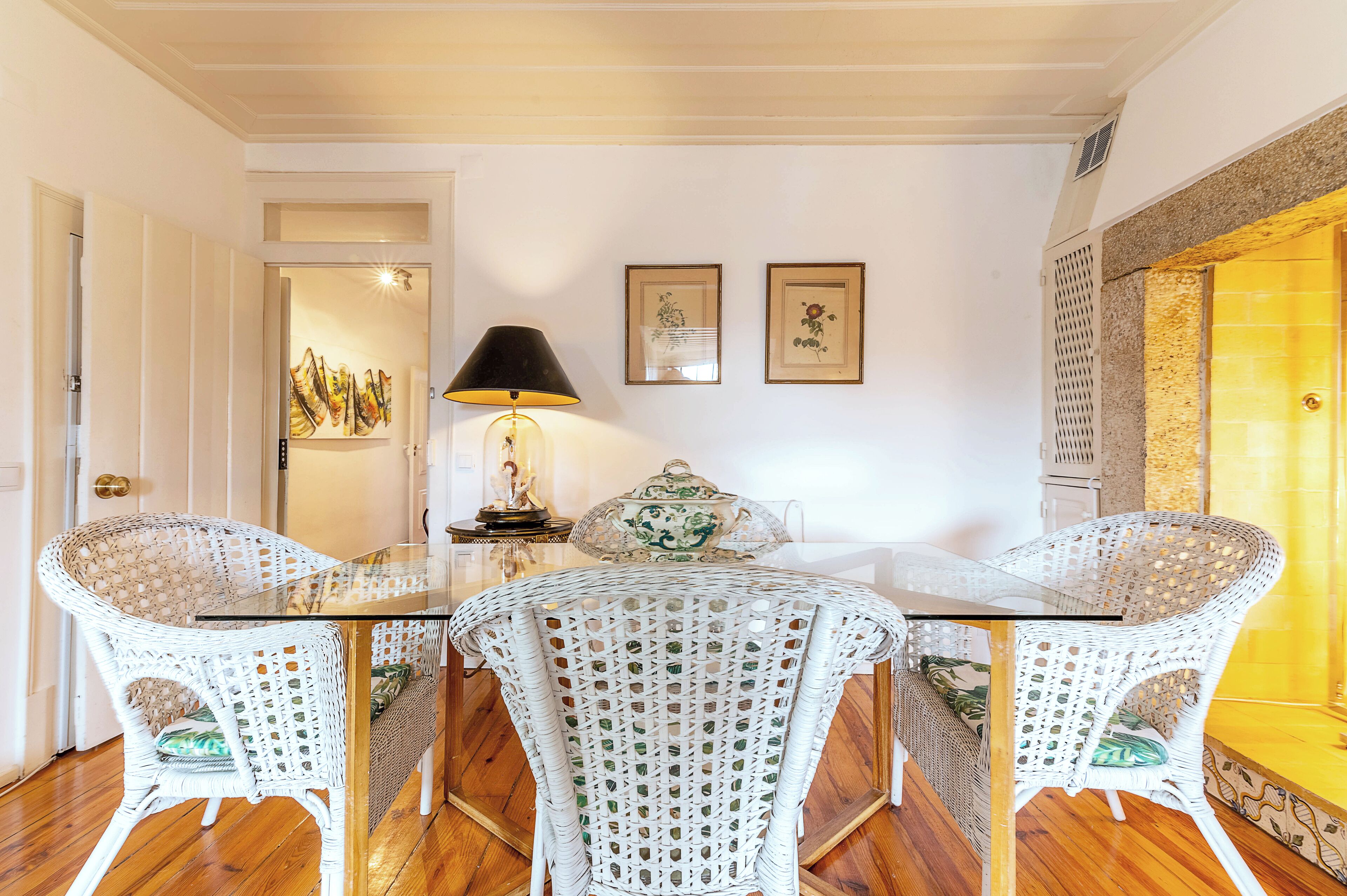A dining area with a glass table, white wicker chairs, a black lamp, and framed pictures on the wall.
